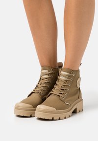 Botines tobilleros marrones con parte superior de lona, punteras de goma y suela con tacos. Presentan ojetes metálicos y una etiqueta de logo en el lateral.