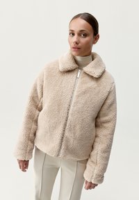 Beżowa kurtka z futra sztucznego z zapięciem na zamek, dużym kołnierzem i przesztywnionymi rękawami. Posiada miękką, teksturowaną powierzchnię oraz luźny krój.