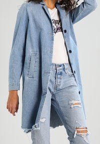 Abrigo de mezclilla azul claro con diseño entallado, seis botones, mangas largas, bolsillos laterales y una parte trasera ligeramente más larga. Se lleva sobre una camiseta gráfica.