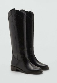 Botas de cuero negras hasta la rodilla con una textura suave, puntas afiladas, costuras decorativas y un tacón bajo en bloque. Presentan un diseño en forma de onda en la parte superior.