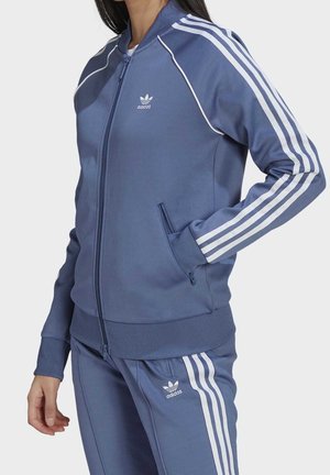 Vrouw draagt blauwe Adidas trainingsjas en broek met witte strepen, staand met één hand in de zak tegen een effen achtergrond.