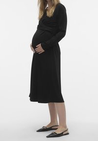 Mujer embarazada con un vestido largo negro y zapatos de tacón bajo negros, sosteniendo su barriga con ambas manos.