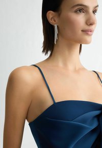 Modré saténové šaty s tenkými ramínky, s jemnými záhyby. Modelka má na sobě dlouhý, třpytivý stříbrný náušnice s kaskádou rhinestones.