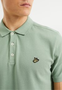 Polo en coton vert clair avec une finition texturée, trois boutons et une broderie d'oiseau noir et jaune sur la poitrine gauche.