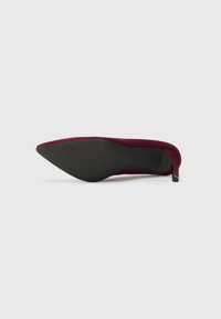 Scarpa con tacco alto color bordeaux, con punta affilata, realizzata in materiale morbido, dotata di una suola nera texturizzata e un tacco sottile nero.