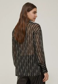 Blusa nera trasparente con maniche lunghe, caratterizzata da un motivo a catena argentata. Presenta un orlo curvo e polsini con bottoni. Tessuto leggero e fluido.