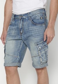 Vaaleansiniset denimit cargo-shortsit, joissa on useita taskuja, hieman kulutettu pinta, taitto helman kohdalla ja nappikiinnitys vyötäröllä.