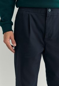 Marineblauwe broek met plooien, gemaakt van een lichtgewicht stof. Voorzien van een elastische tailleband en een knoopsluiting, met een gladde textuur.