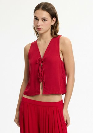 Joven mujer vistiendo una blusa roja sin mangas con lazos en la parte frontal y una falda roja plisada a juego, de pie frente a un fondo blanco.