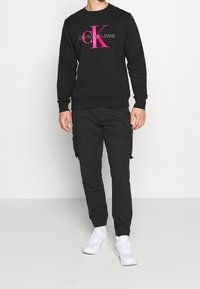 Sudadera negra con el logo y texto "CK" en rosa, combinada con pantalones cargo negros y zapatillas blancas. Tejido suave, puños y dobladillo acanalados.