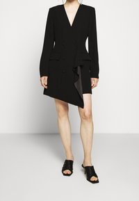 Robe-veste noire à double boutonnage avec un décolleté en V profond, manches longues et ourlet asymétrique agrémenté d'un détail à volants. Portée avec des sandales plates noires.