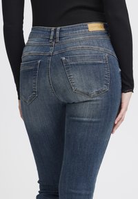 Tamnoplave traperice uskog kroja s pet džepova, vidljivim šavovima i etiketom na pojasu. Tekstura je glatka i blago istrošena.