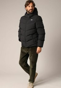 Doudoune noire avec fermeture éclair et capuche, présentant des sections matelassées et des poches latérales. Associée à un pantalon en velours côtelé olive et des chaussures beiges.