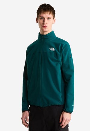 Ung mand iført en teal North Face jakke med fuld lynlås og høj krave samt sorte bukser, stående mod en hvid baggrund.