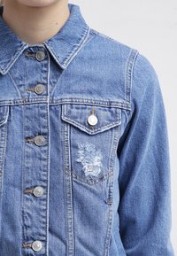 Person som bär en ljusblå denimjacka med metallknappar och en sliten lapp på övre vänstra bröstfickan.