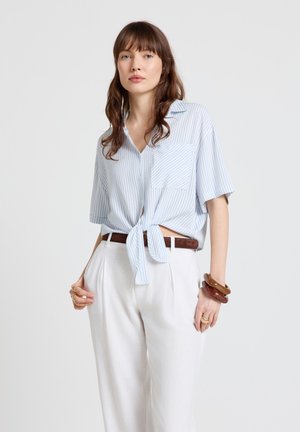 Woman wearing light blue striped short-sleeve shirt tied at waist, white high-waisted pants, brown belt, and chunky bracelets.