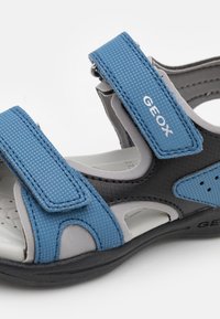 Sandalia abierta en azul y gris con dos correas ajustables de Velcro, parte superior de material sintético texturizado y lados perforados para mayor transpirabilidad.