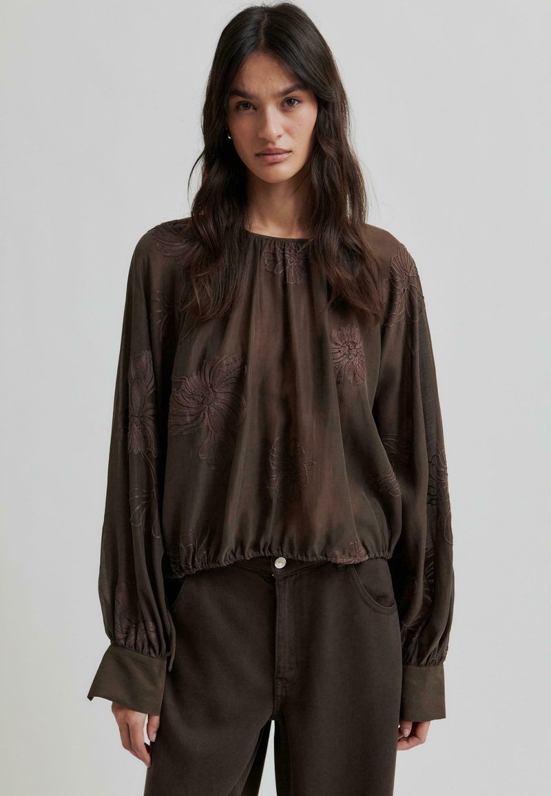 Blouse légère brun avec broderies florales, manches longues avec larges poignets et taille froncée. Associée à un pantalon sombre.