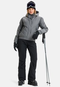 Giacca isolata grigia con cappuccio, pantaloni neri, guanti neri, occhiali da sci e sci; progettato per sport invernali, con uno stile aderente.