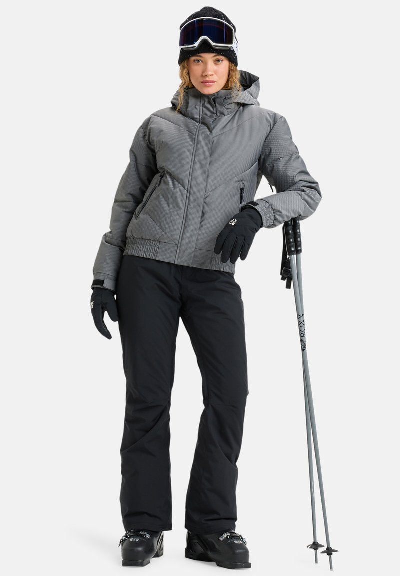 Giacca isolata grigia con cappuccio, pantaloni neri, guanti neri, occhiali da sci e sci; progettato per sport invernali, con uno stile aderente.