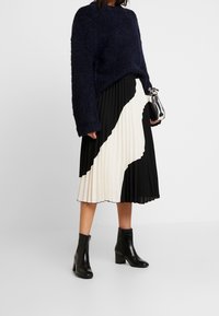 Osoba ubrana w granatowy sweter i czarno-białą plisowaną spódnicę midi, trzymająca srebrną torbę, w czarnych botkach za kostkę, na białym tle.