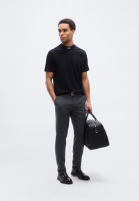 Άνδρας που φοράει μαύρο κοντομάνικο πουκάμισο και γκρι παντελόνι κρατάει μαύρη δερμάτινη τσάντα, στέκεται μπροστά σε απλό φωτεινό φόντο.