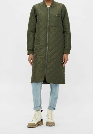 Manteau matelassé long vert olive avec fermeture éclair à l'avant, poche poitrine, porté sur un jean bleu clair retroussé et des bottines beiges à lacets.