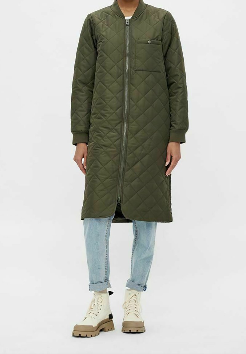 Manteau matelassé long vert olive avec fermeture éclair à l'avant, poche poitrine, porté sur un jean bleu clair retroussé et des bottines beiges à lacets.