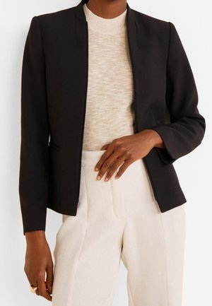 Frau trägt einen schwarzen, maßgeschneiderten Blazer über einem beigen Oberteil, kombiniert mit hoch taillierten, hellen Hosen, Hand auf der Taille ruhend.