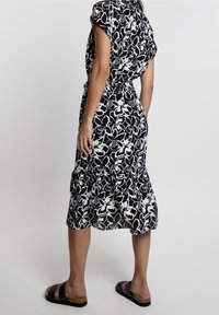 Schwarz-weißes, blumengemustertes Kleid mit kurzen Ärmeln, tailliertem Schnitt und rüschendem Saum, kombiniert mit schwarzen Rutsch-Sandalen.