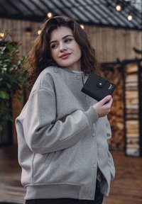 Black wallet held in a hand, featuring a snap closure and logo. The wearer is in a grey, oversized jacket with ribbed cuffs and a smooth texture.
