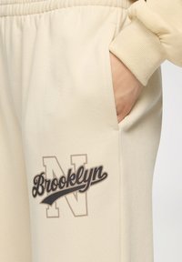 Jasnokremowe dresy wykonane z miękkiego materiału, z boczną kieszenią i ciemną grafiką z napisem "Brooklyn" oraz dużym logo "N".