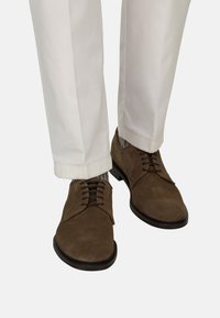 Chaussures habillées en daim marron à lacets portées avec un pantalon blanc et des chaussettes à motifs, visibles de mi-mollet vers le bas sur un fond neutre.