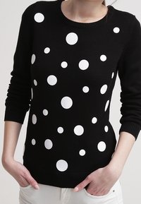 Czarny sweter w białe groszki, z prążkowanym dekoltem i mankietami, dopasowany krój oraz teksturowana dzianina. Ręce ułożone na talii.