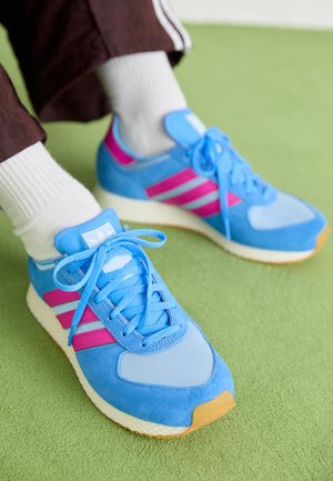 Jasnoniebieskie i różowe sneakersy z białymi skarpetkami na zielonym dywanie, noszone przez osobę w brązowych spodniach z białymi paskami.