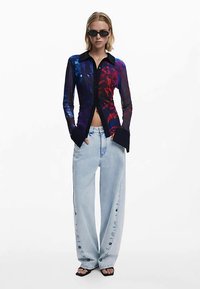 Langärmeliges durchsichtiges Hemd mit blauem und rotem Blumenmuster, schwarzem Kragen und Manschetten, kombiniert mit hellblauen, weiten Jeans mit Knopfdetails.