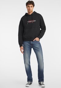 Svart hettegenser med rød "GUESS JEANS" logo, kengurulomme, justerbar hette med snor, kombinert med lyseblå denimjeans og svarte sneakers.