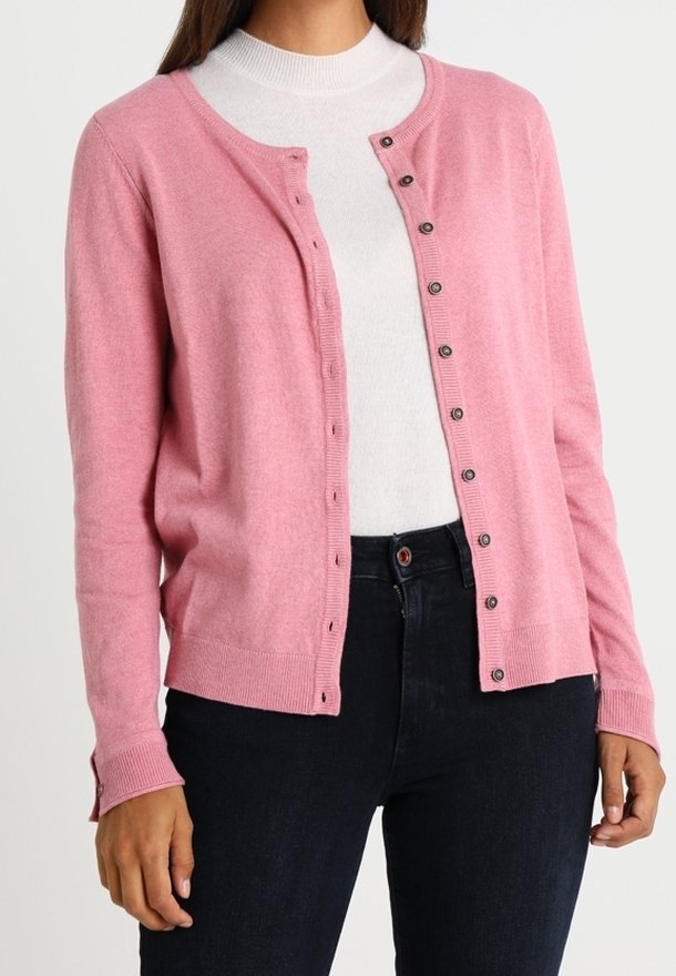 Cardigan rose clair en tissu doux, avec un col rond, une fermeture à boutons, et des poignets et une base côtelés. Porté sur un haut blanc.