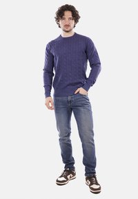 Maglione lavorato a maglia viola con motivo a trecce, collo rotondo, maniche lunghe, abbinato a jeans blu e sneakers marroni. Texture liscia.