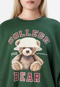 Primo piano di una felpa verde con un orsetto che indossa un cappello bianco di paglia e la scritta "COLLEGE BEAR" in lettere rosse.