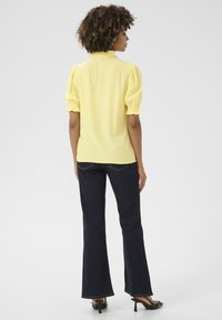 Blusa gialla a maniche corte con maniche a sbuffo e colletto sul retro, abbinata a jeans a zampa d'elefante blu scuro e sandali neri con tacco.