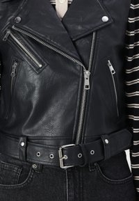 Schwarze Leder-Motorradjacke mit silbernen Reißverschlüssen, diagonalen Taschen, einem belüfteten Bund und einer strukturierten Oberfläche. Gestreiftes Hemd darunter sichtbar.
