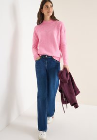 Różowy sweter wykonany na drutach z ribbowymi mankietami, wzorzysty; niebieskie spodnie z szerokimi nogawkami; trzyma głęboki fioletowy hoodie; białe sneakersy.