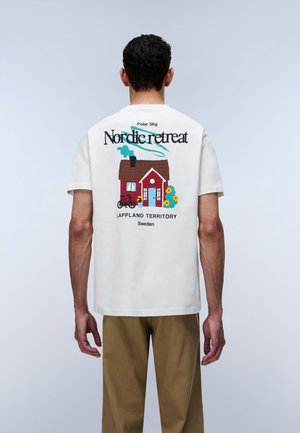 Man wearing white T-shirt with colorful graphic of red house, bicycle, flowers, and text "Nordic retreat Lappland Territory Sweden" on back.