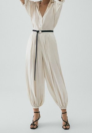 Woman wearing a beige buttoned jumpsuit with a black belt, gathered pants at ankles, and black strappy high-heeled sandals.