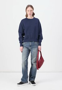 Marinblå sweatshirt med rund halsringning, löst sittande och ribbad nederkant; ljusblå jeans; röd väska; svarta sneakers.