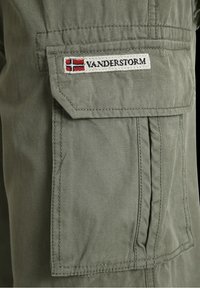 Zielone oliwkowe spodnie cargo z kieszenią naszywaną, ozdobioną haftowaną etykietą "Vanderstorm" w kolorach czerwonym, białym i niebieskim. Wytrzymały materiał z widocznymi ściegami.