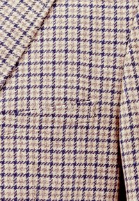 Beiger Stoff mit einem Hahnentrittmuster in Dunkelblau. Die Textur ist gewebt mit einem leichten Glanz und verfügt über ein kleines Taschendetail auf der Vorderseite.