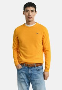 Leuchtend gelb gestrickter Pullover mit rundem Halsauschnitt, lockerer Passform und dezentem gesticktem Logo. Kombiniert mit hellblauen Jeans und einem braunen Gürtel.