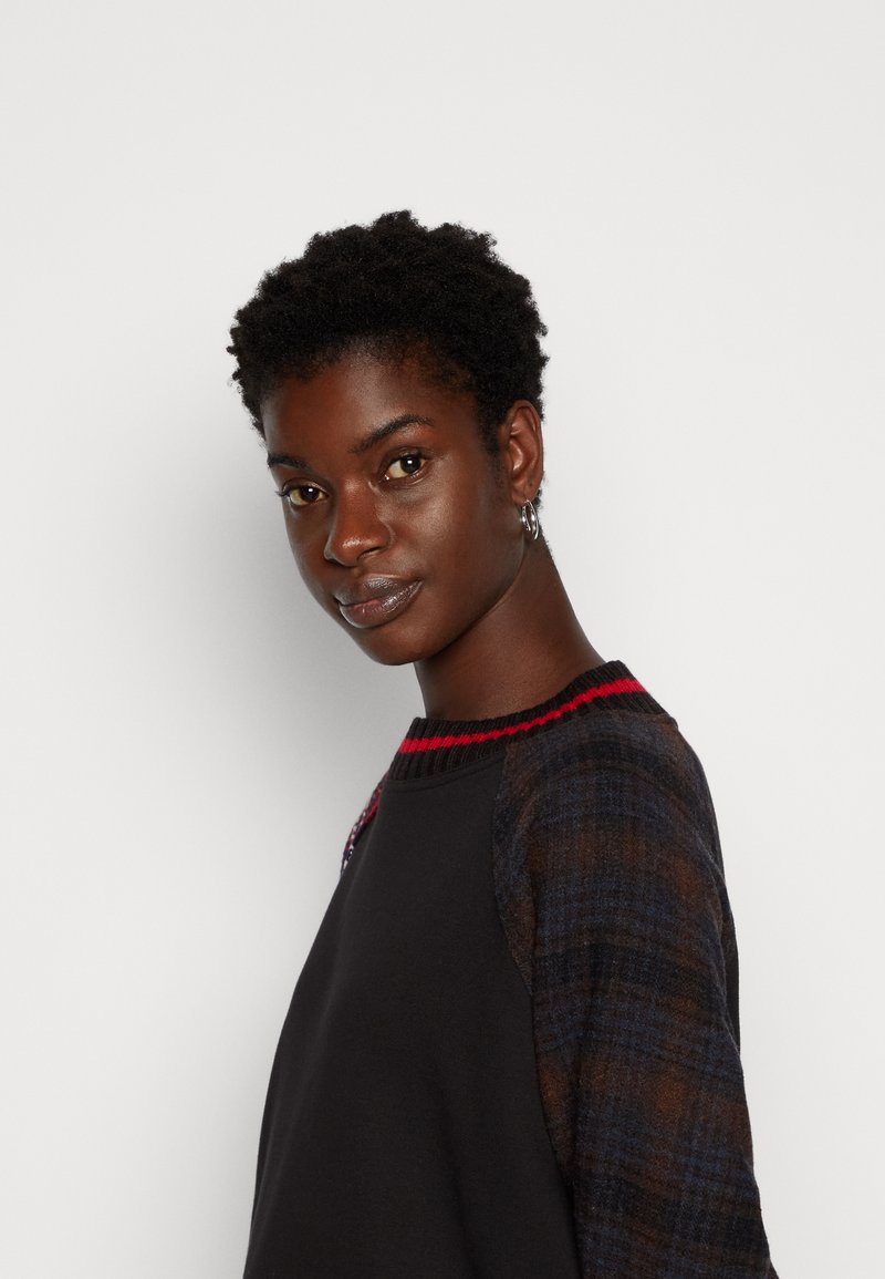 Czarny sweter z rękawami z materiału w kratę, czerwonym ściągaczem przy szyi i detale logo na klatce piersiowej. Gładka struktura z różnorodnymi wzorami materiałowymi.